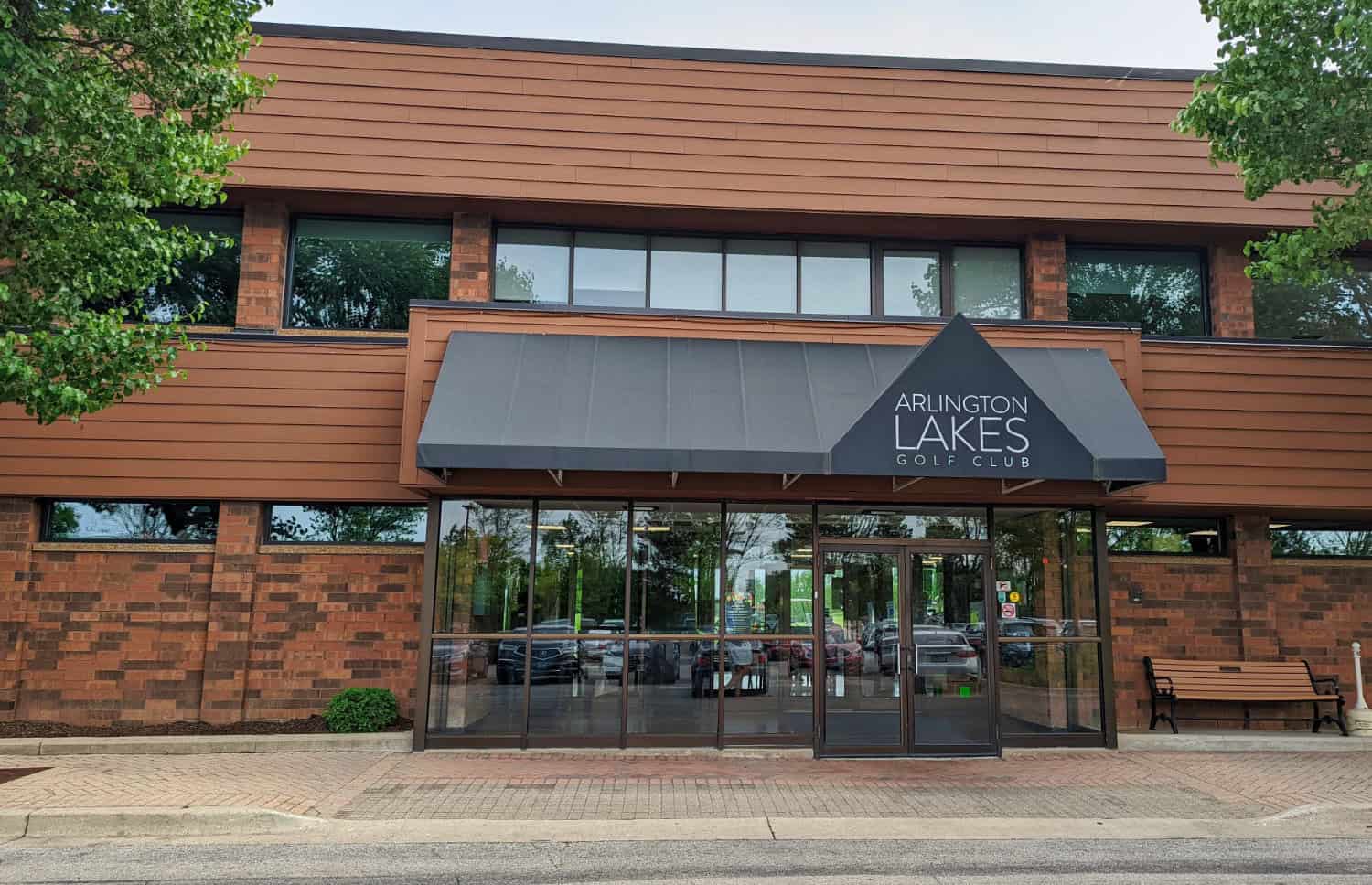 The entrance to Arlington Lakes Golf Club, showing a brick building with large windows, a black awning, and glass doors.