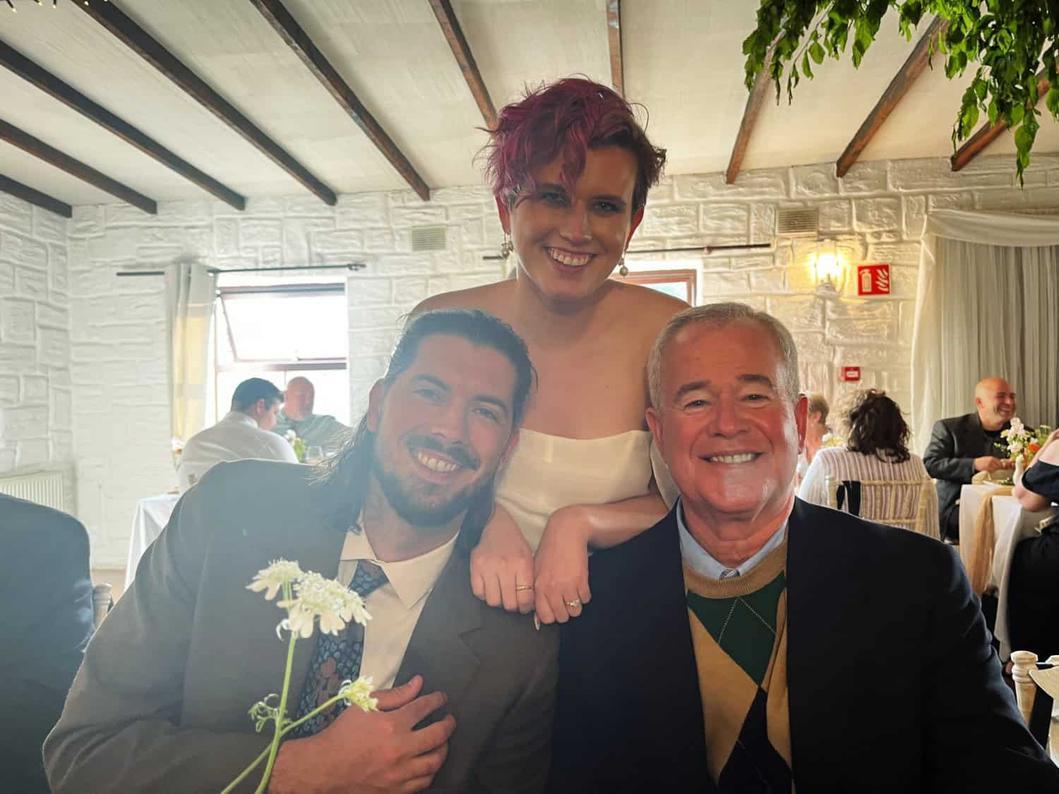 Three people pose and smile at a table during a formal event, with other guests visible in the background.