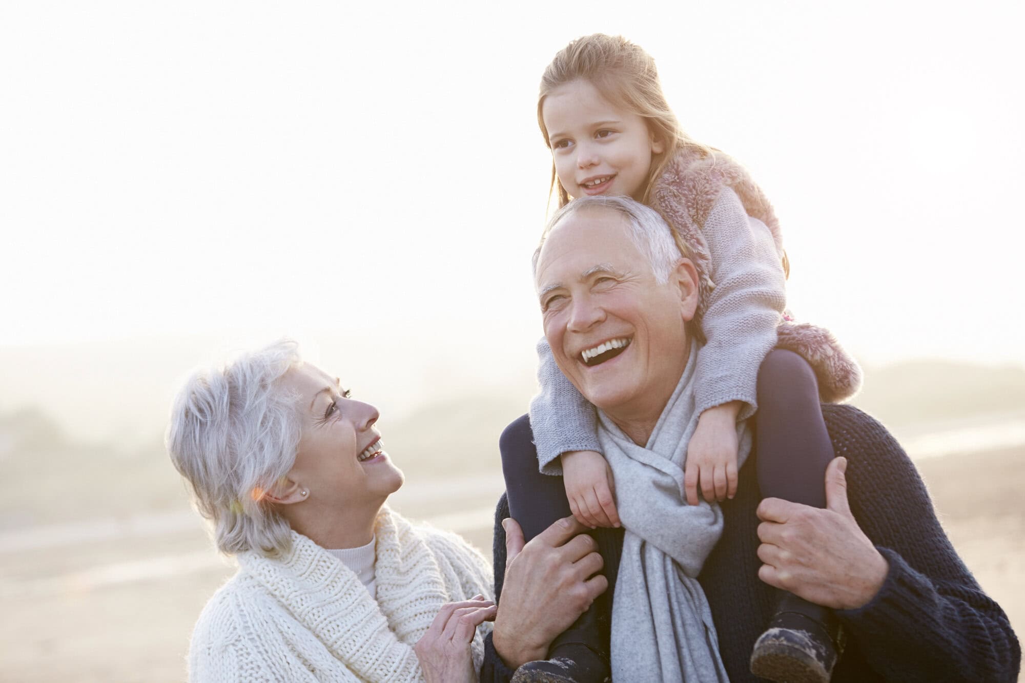 Older couple and young girl outdoors; the man carries the girl on his shoulders while the woman smiles beside them, all dressed warmly on a bright, possibly chilly day.